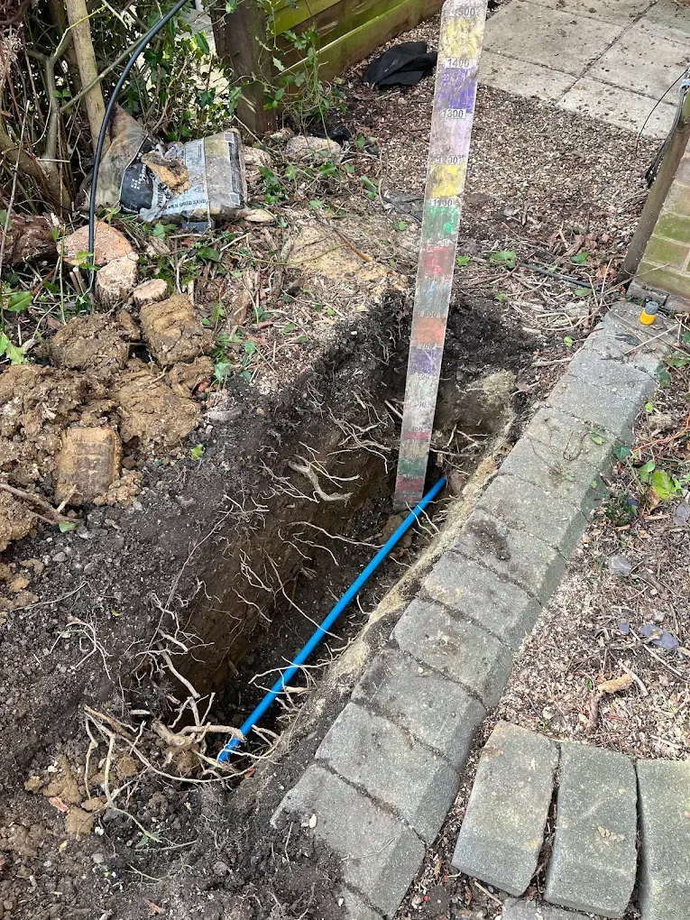 A trench along a garden border beside a low brick wall, new blue pipe laid along the bottom