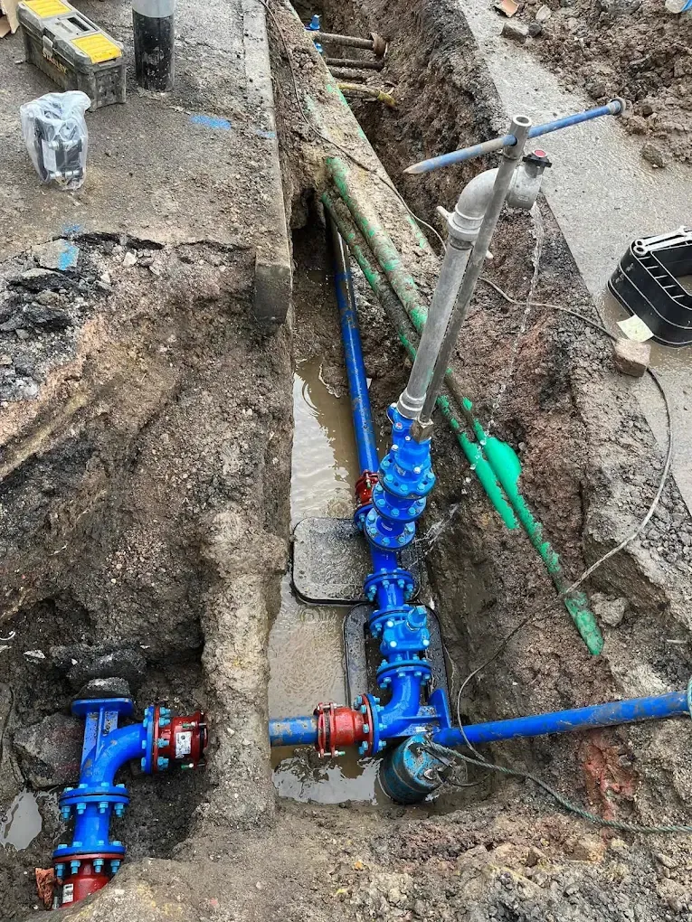 Blue valves and red fittings connected in a trench along a pavement edge, with new green and blue pipes running off