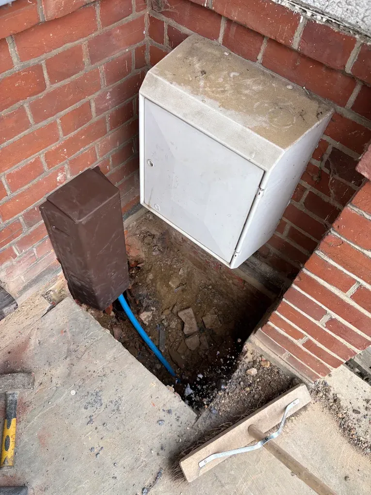 An excavation in the corner by a wall-mounted meter cabinet, with new blue pipe running up to it