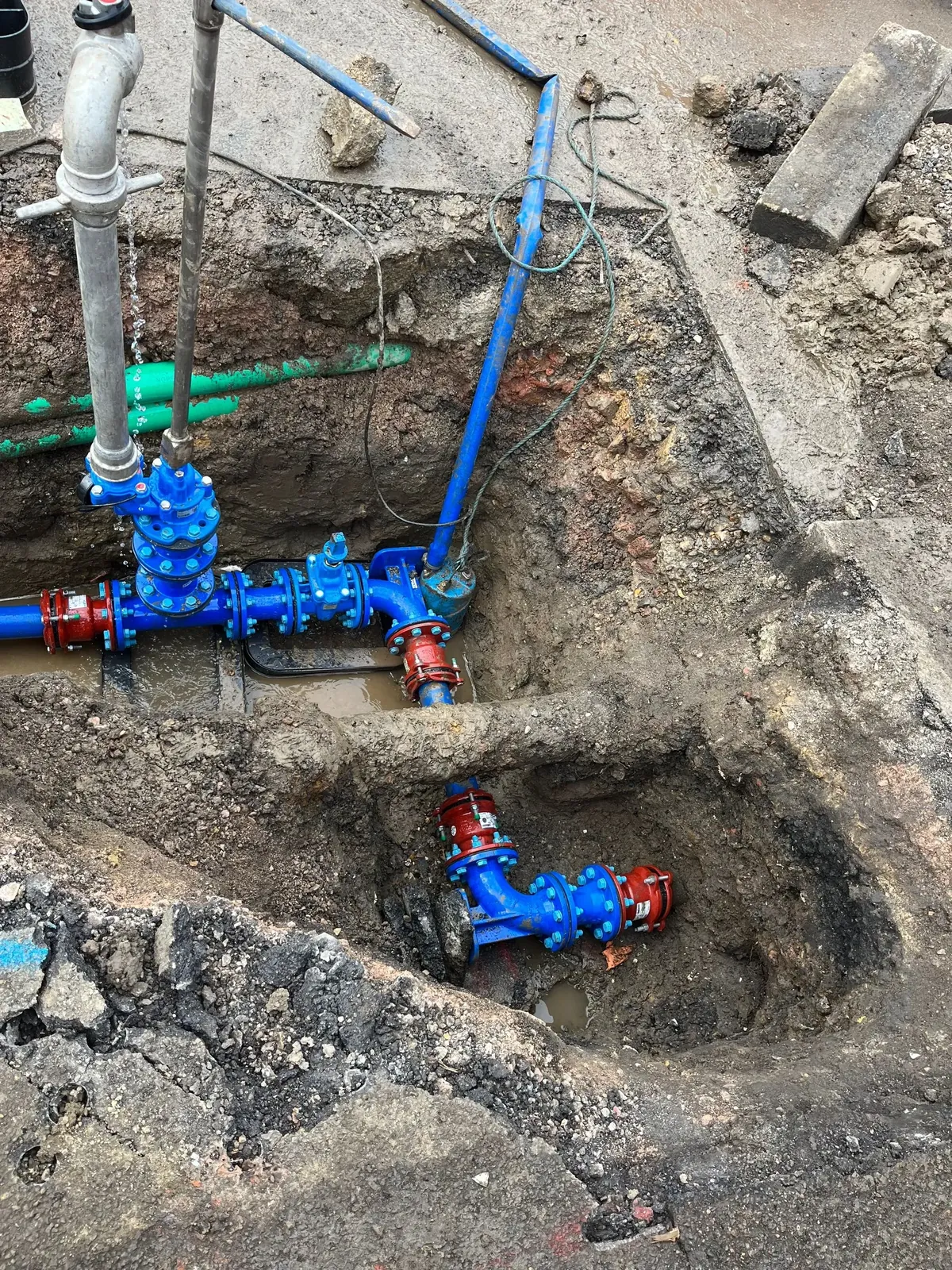 Two excavations at the edge of a pavement with new blue valves and supply pipes connected beside an older grey pipe