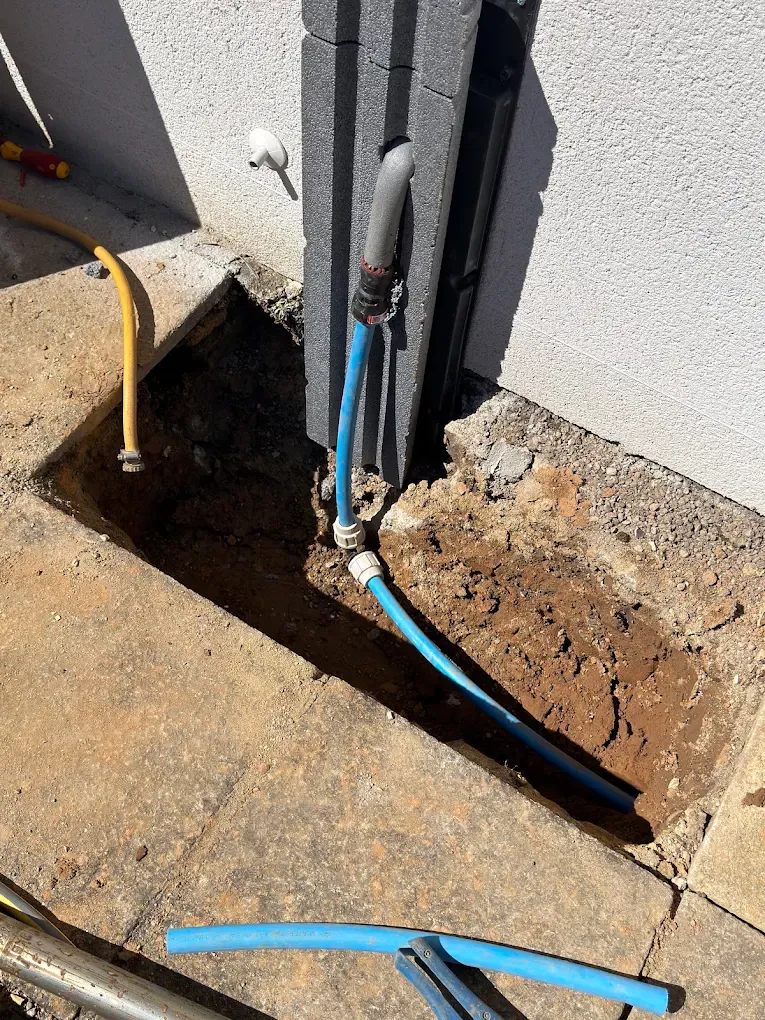 New blue supply pipe rising out of a pit beside a rendered house wall, ready to enter the building