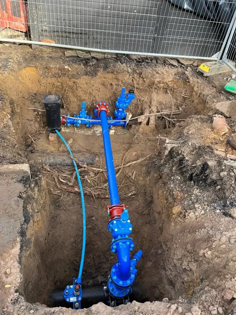 A deep excavation behind site fencing with several new blue supply pipes and valves branching from one connection