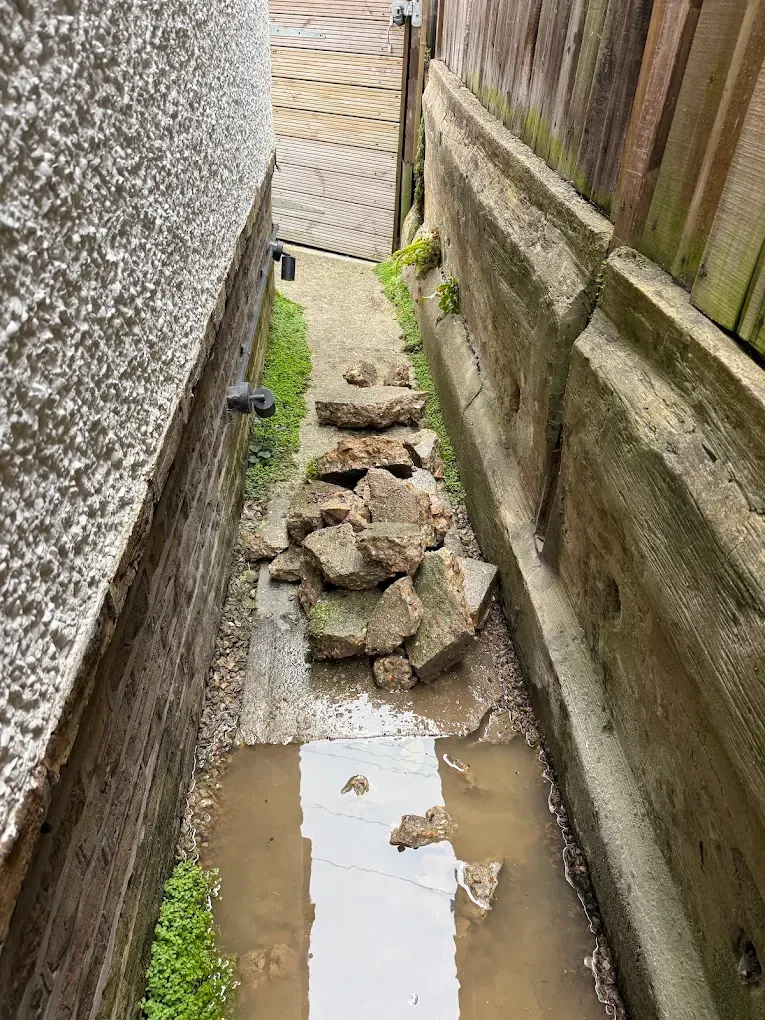 A narrow side passage with water pooled on the path and broken concrete stacked against the wall