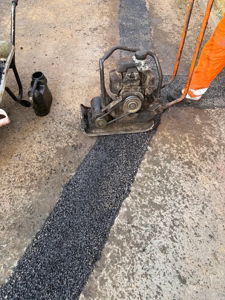 A plate compactor rolling a fresh strip of tarmac into a pavement trench